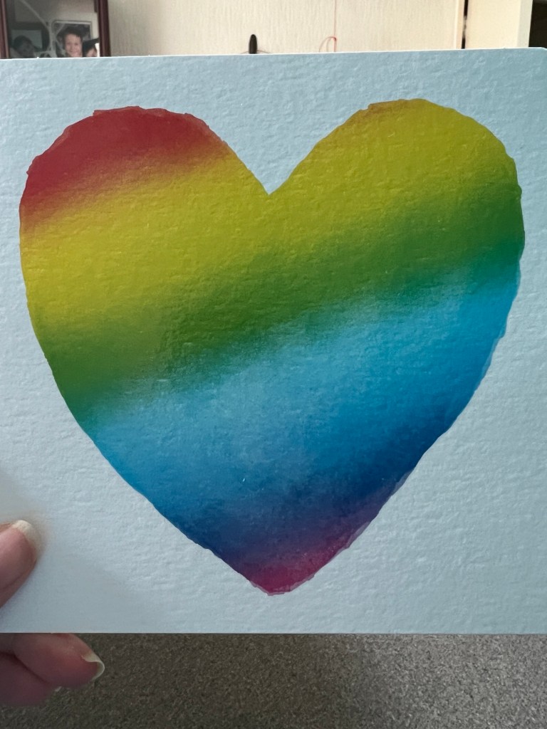 Square card with a large heart made as if someone used watercolour paints in rainbow colours to make wide diagonal stripes through a stencil from red top left in order to violet at the bottom tip