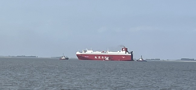The large red Kess ship on the grey water with 3 smaller ships (one each in front, behind, and beside it)