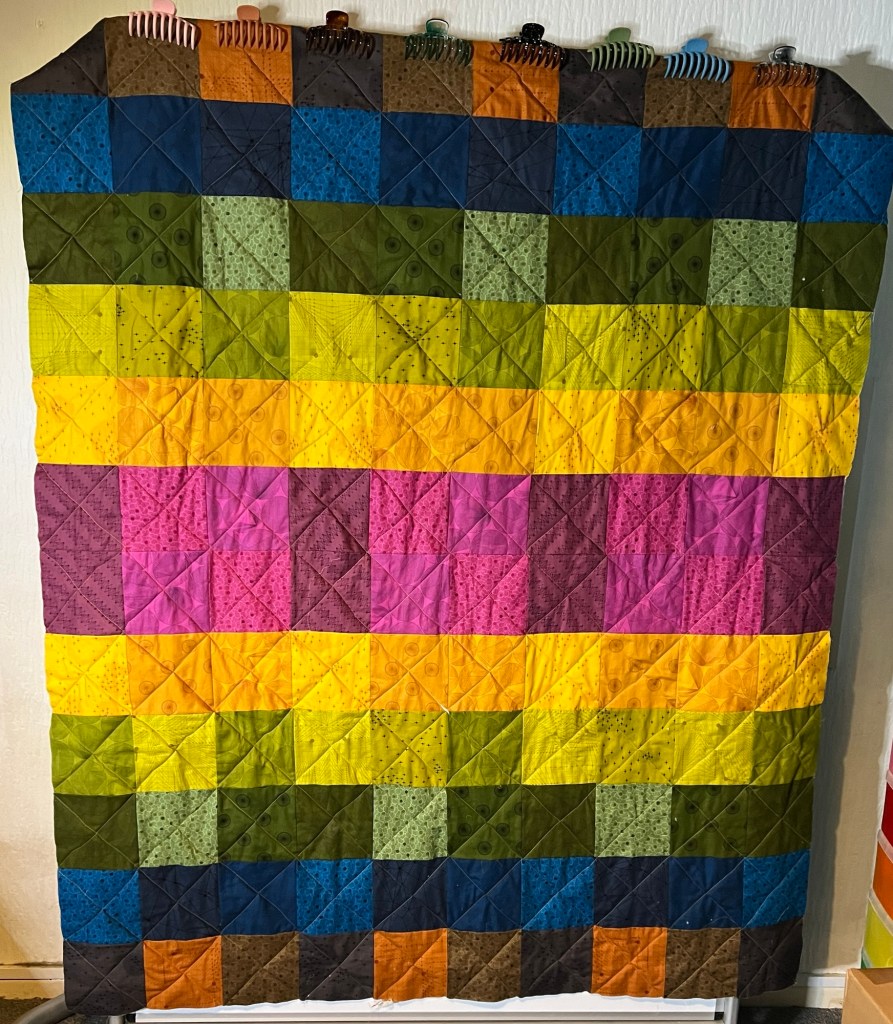 After Wrestling While Sewing (aka quilting) with a simple diagonal cross hatch.

The image shows a colorful quilt made up of square patches arranged in a grid pattern. The quilt features a variety of colors, including shades of blue, green, yellow, orange, pink, and brown. Each square patch has a subtle pattern or texture, adding visual interest to the quilt. The quilt is hanging vertically, and the top edge is clipped with several hair clips in different colors, which are used to hold it in place. The overall appearance is vibrant and visually striking, with a harmonious blend of colors.