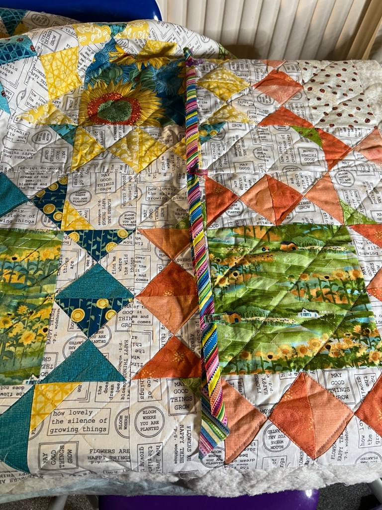 A colourful quilt with a patchwork design. The quilt features various patterns and colors, including blue, yellow, orange, and green. Some sections have floral designs, such as sunflowers, and there are also sections with text. The text includes phrases like "how lovely the silence of growing things," "may good things grow," "bloom where you are planted," and "flowers are happy things." There is a multicoloured, striped binding laid across the quilt so I could see if it looked right.