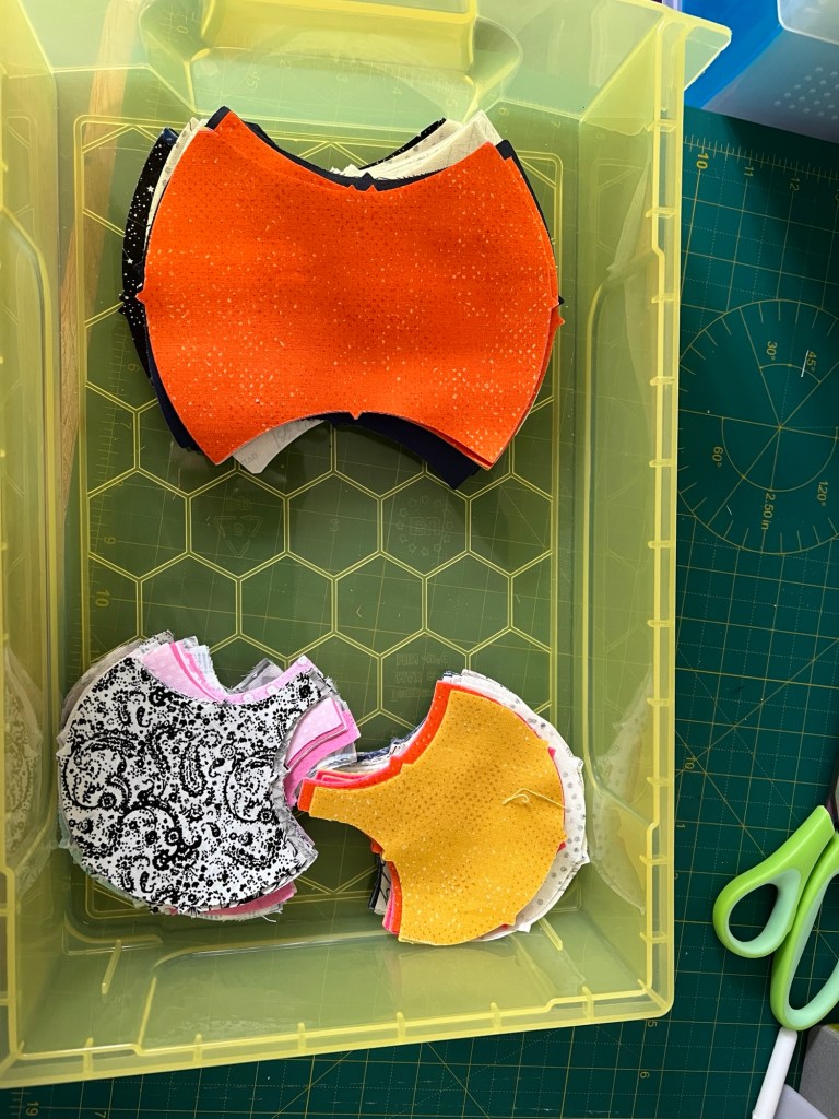 A yellow plastic container holds several stacks of fabric pieces cut into specific shapes. The top stack of apple cores has bright orange fabric with small white speckles. Below it, there are 2 stacks of clamshells which contain fabric with a black and white paisley pattern, pink fabric, and a yellow fabric with small white speckles. The container is placed on a green cutting mat with measurement markings, and a pair of green-handled scissors is visible nearby.
