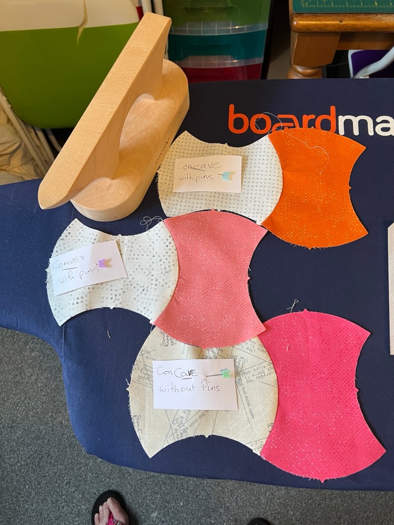 A wooden seam presser tool is placed on a dark blue surface with the word "board" visible in orange and white letters. Below the tool, there are several pieces of fabric in different colors and patterns, cut into curved shapes. The fabrics include white with small dots, pink, orange, and a pattern resembling a sewing pattern. Each piece of fabric has a small note pinned to it. The notes read:

- "Concave with pins" on a white fabric with small dots.
- "Convex with pins" on another white fabric with small dots.
- "Concave without pins" on a fabric with a sewing pattern design.
