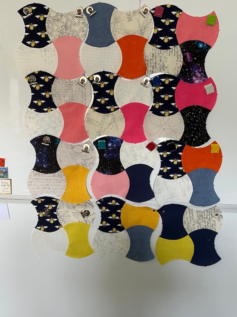 A collection of fabric pieces arranged in a grid pattern, each with a unique design. The fabric pieces are shaped like apple cores and are clipped to a white surface with small metal clips. The designs include bees on a dark background, pink and orange solid colors, a galaxy pattern, and various prints with handwritten text and geometric patterns. Some pieces have a dotted or speckled design. The arrangement creates a colorful and visually interesting patchwork effect.