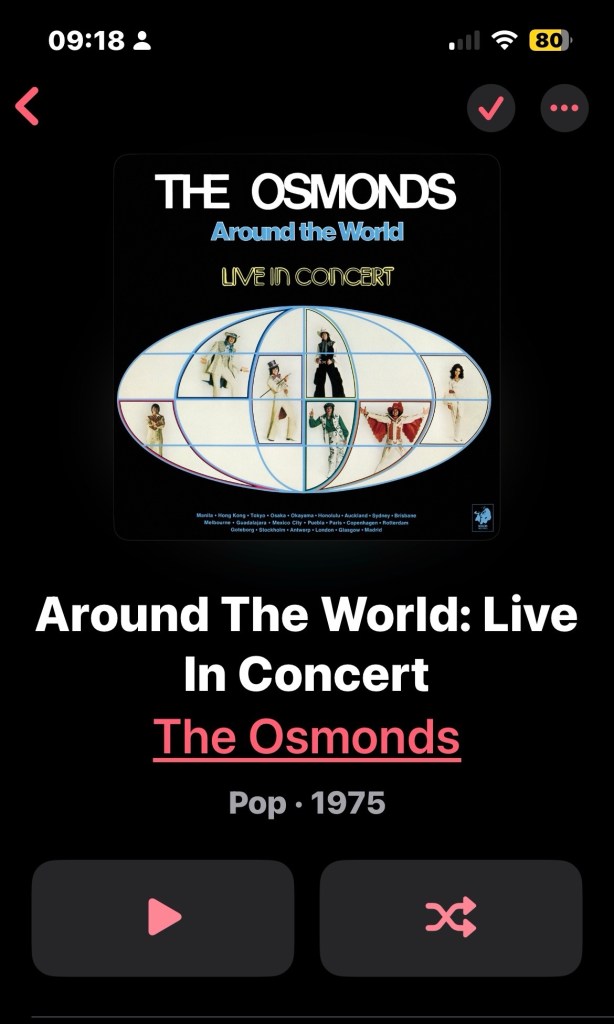 Album cover for "Around The World: Live In Concert" by The Osmonds. The cover features a black background with a stylized globe in the center, divided into sections. Inside each section, there are photos of the band members in colorful stage outfits, each posing differently. The text at the top reads "THE OSMONDS" in large white letters, "Around the World" in blue, and "LIVE IN CONCERT" in yellow. Below the globe, there is a list of cities in small blue text. The app interface shows the album title in large white text, the artist name "The Osmonds" in pink, and the genre and year "Pop · 1975" in gray. At the bottom, there are two large buttons: a pink play button on the left and a pink shuffle button on the right. The time at the top left is 09:18, and the battery indicator at the top right shows 80%.