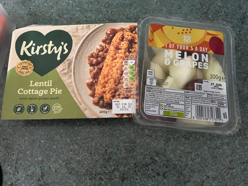 A food package and a fruit container on a dark green countertop. The food package is labeled "Kirsty's Lentil Cottage Pie with sweet potato mash." It has a green and beige design with a heart-shaped logo and icons indicating it is gluten-free, dairy-free, vegan, and contains 236 calories per serving. The package shows an image of the cottage pie with lentils and sweet potato mash on a plate. The weight is 400 grams, and there is a use-by date sticker that reads "30 JUL 25."

Next to it is a clear plastic container labeled "1 of your 5 a day Melon & Grapes" from Co-op. The container holds 300 grams of melon and grapes, with a use-by date of "27 JUN." The label includes nutritional information, ingredients, and recycling instructions. The melon pieces inside are light in color, and the grapes are not clearly visible from this angle.