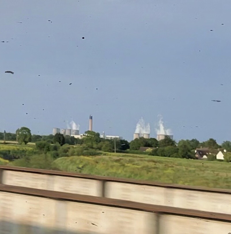 Power station in the distance, with several large cooling towers emitting white steam into the sky. The scene is set in a rural area with green fields, scattered trees, and a few houses visible near the right side of the image. In the foreground, there is a blurred wall as seen from the moving train. The sky is clear and blue, with some dark spots and marks on the image from a dirty window. 