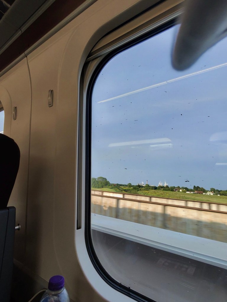 Train window view. The photo is taken from inside a train carriage, looking out through a large window. The interior is modern, with smooth beige walls and a dark seat partially visible on the left. Outside the window, there is a blur of a bridge or barrier close to the train, and beyond that, a green landscape with trees and a few distant buildings under a blue sky. The window has some smudges and spots on it. At the bottom left corner, there is a plastic bottle with a purple cap resting on a small table or ledge. The overall scene suggests the train is moving through the countryside.