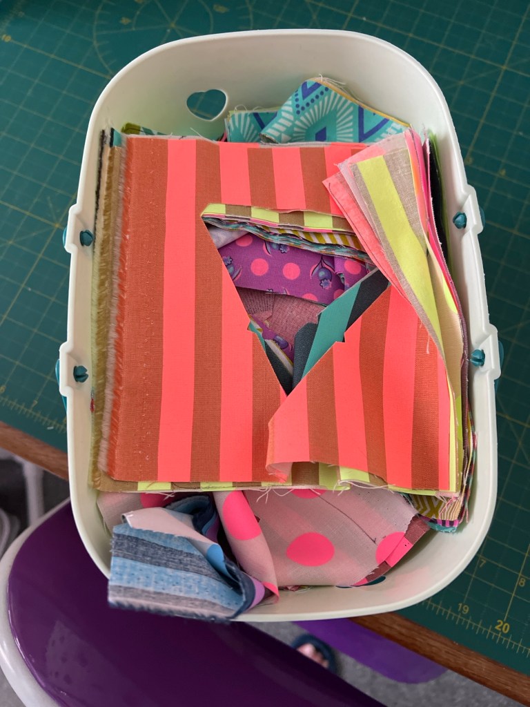 A container filled with colourful fabric scraps. The fabrics have various patterns, including bright pink and orange stripes, pink polka dots on gray, purple with small designs, and some geometric shapes. The pieces are layered and crumpled together. 