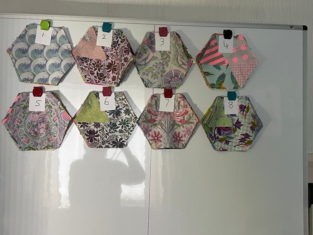 Eight stacks of hexagon-shaped fabric pieces, each with a different pattern and color, are attached to a whiteboard. Each stack is held in place by a colored magnet and labeled with a small piece of paper numbered from 1 to 8. The stacks are arranged in two rows of four. The patterns on the fabrics include waves, florals, birds, and geometric shapes in various pastel and bright colors. The whiteboard is otherwise empty except for the shadow of a person taking the photo.