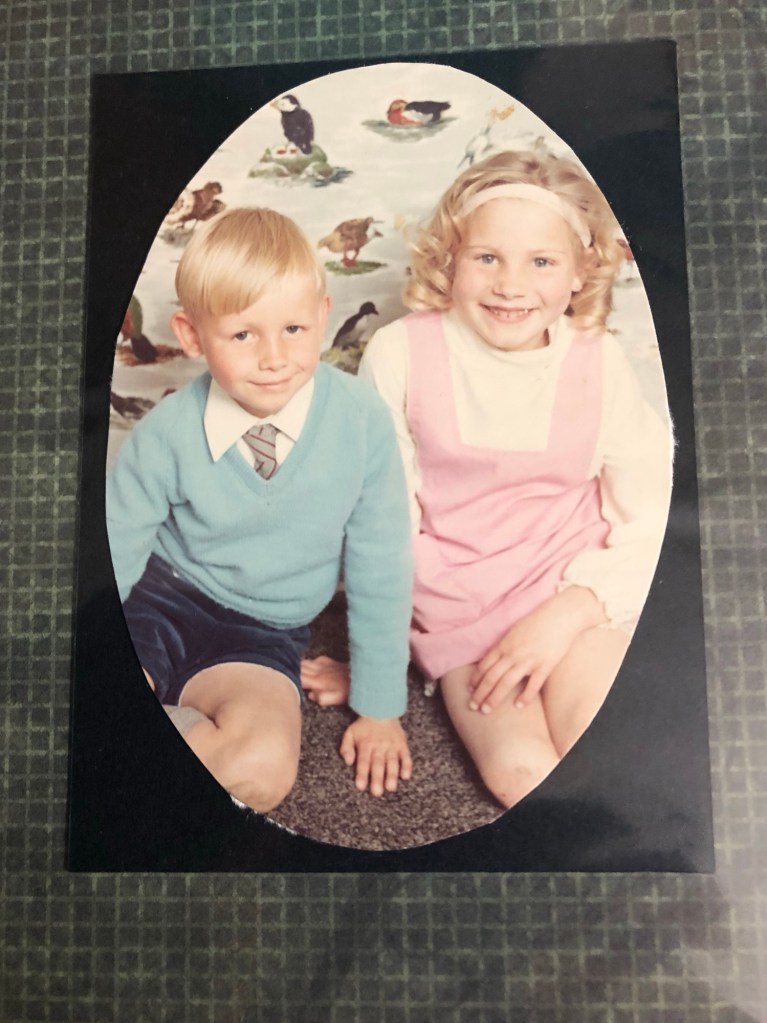 A vintage photograph of two young children sitting side by side. The boy on the left has short blonde hair and is wearing a light blue sweater over a white collared shirt with a striped tie, along with dark shorts. The girl on the right has curly blonde hair with a headband and is wearing a pink pinafore dress over a white long-sleeved shirt. Both children are smiling. The background features a wallpaper with illustrations of various birds. The photo has an oval shape and is mounted on a dark background.
