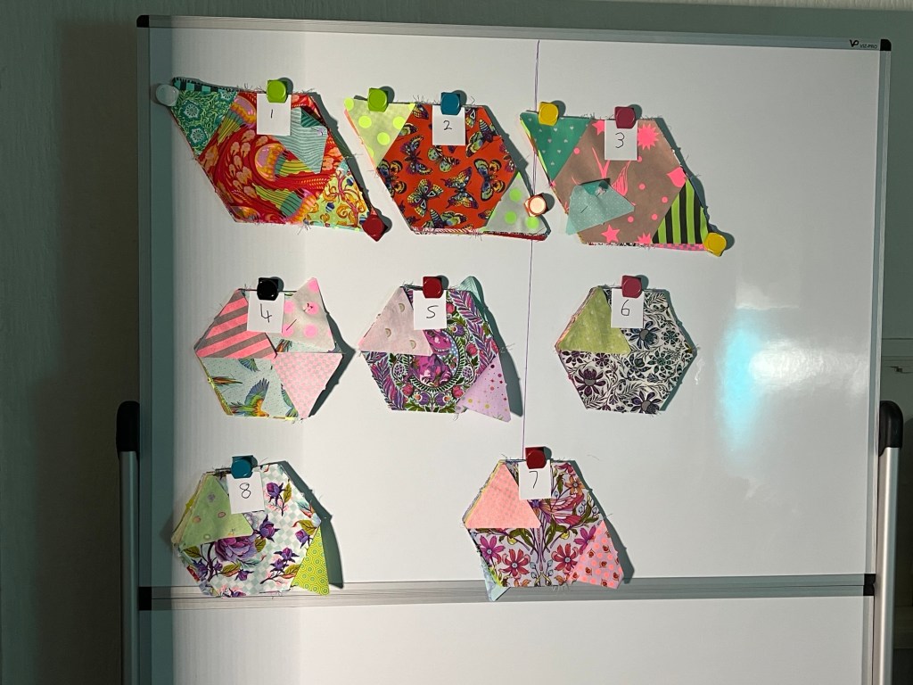 Whiteboard with stacks of colourful fabric diamonds made from a hexagon and 2 equilateral triangles. Each diamond is made from multiple pieces of patterned fabric and has a small paper label with a number from 1 to 8. The numbers are handwritten and each label is held in place by a different colored magnet. The top row has three stacks of diamonds that have been pressed in and the five stacks in the bottom two rows have not yet been pressed. The fabrics feature a variety of patterns, including florals, birds, stripes, and abstract designs in bright colors like pink, green, orange, and purple. The whiteboard is standing on a frame, and the lighting creates some shadows on the left side.