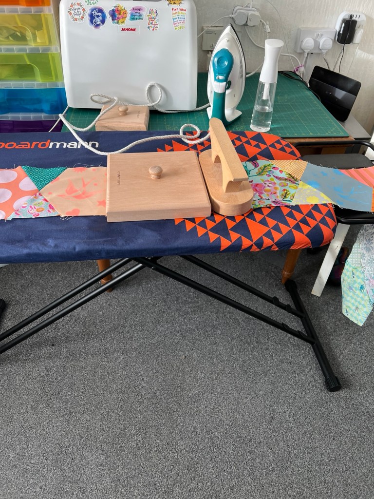 Sewing and ironing station. There is an ironing board covered with a colourful geometric and floral fabric, and on top of it are wooden tools, including a tailor’s clapper and two wooden pressing blocks. There is a strip of patchwork fabric laid out on the board. In the background, there is a white sewing machine with stickers on it, a steam iron, a spray bottle, and a green cutting mat. To the left, there are colourful plastic drawers for storage. The floor is carpeted in grey. This setup suggests a workspace for quilting or sewing projects.