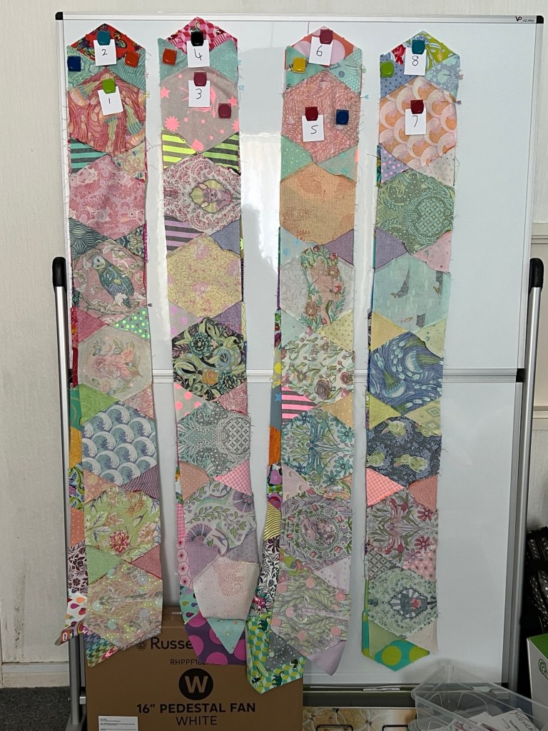 Four long, narrow strips of patchwork fabric, each made from hexagonal pieces in various pastel and bright patterns, are hanging vertically on a whiteboard. Each strip is secured at the top with colorful magnets and labeled with small white cards numbered 1 to 8. The strips are arranged side by side, and each one features a unique combination of floral, geometric, and abstract prints.