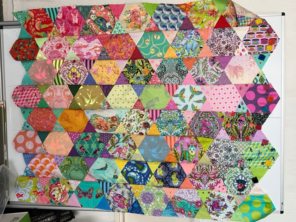 Colourful vs patchwork quilt top made of hexagon and triangle shaped fabric pieces. Each piece features a different vibrant pattern, including florals, animals, birds, geometric shapes, and abstract designs in a wide range of colours such as pink, green, blue, yellow, orange, and purple. The hexagons and triangles are arranged in rows, and the quilt top is held in place on a white surface with small clips at the edges. The overall effect is lively and visually engaging, with a mix of whimsical and traditional motifs.