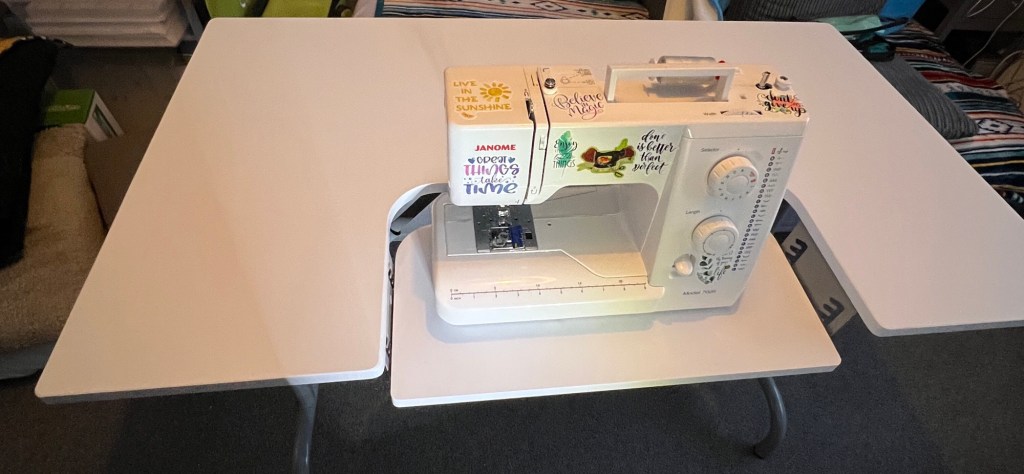 White sewing machine set on a large folding table. The table has two side panels that fold out, creating extra working space on either side of the sewing machine. The sewing machine is decorated with several colourful stickers that include positive messages such as "Live in the Sunshine", "Great things take time", "Believe in magic", and "Don't give up". The sewing area is clean and organized, and in the background, is a seating area with a striped blanket and several cushions. Some boxes and items are partially visible on the left side. The environment looks like a cosy craft or sewing room.