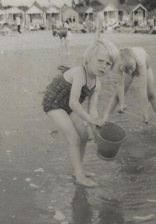 Black-and-white photograph of a young child at the beach. The child (5 year old me), who has short light-colored hair and is wearing a sleeveless swimsuit, is crouched down at the water's edge holding a bucket. Another child is close by, bending down and reaching toward the water. In the background, there are rows of beach huts and some people visible further away. The scene suggests a nostalgic, possibly mid-20th century seaside holiday.
(1967 Walton on the Naze)