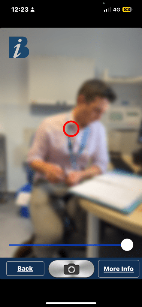 A blurry image of a person sitting at a desk

The image is very blurry, making it difficult to see details, but it shows a person sitting at a desk, writing or working with papers. There is a red circle indicating the centre of the image, and the person seems to be wearing a lanyard. The top of the image has a blue information icon with the letter "i" on it, as well as mobile phone status details (12:23 time, 4G signal, battery at 83%). At the bottom, there are buttons labeled "Back", a camera icon, and "More Info", suggesting this is a screenshot from an app, possibly for identifying or capturing information. The setting looks like an office or clinical environment based on the visible furniture and equipment.