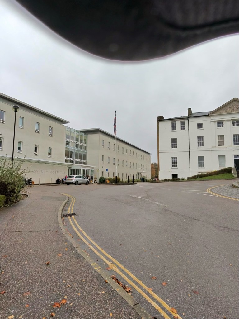 Hospital complex on a cloudy day, with two large white buildings. The building on the left has a modern entrance with glass doors and the words "COUNTY HOSPITAL" partially visible. A UK flag is flying from a flagpole near the entrance. Several cars and some people are near the entrance, mostly on the left side of the image. There’s a roundabout in front of the buildings, marked with double yellow lines along the curb. Leaves are scattered on the ground. The top edge of the photo has a large dark shape of my cap brim, partially covering the upper portion of the image. The sky is overcast and grey.