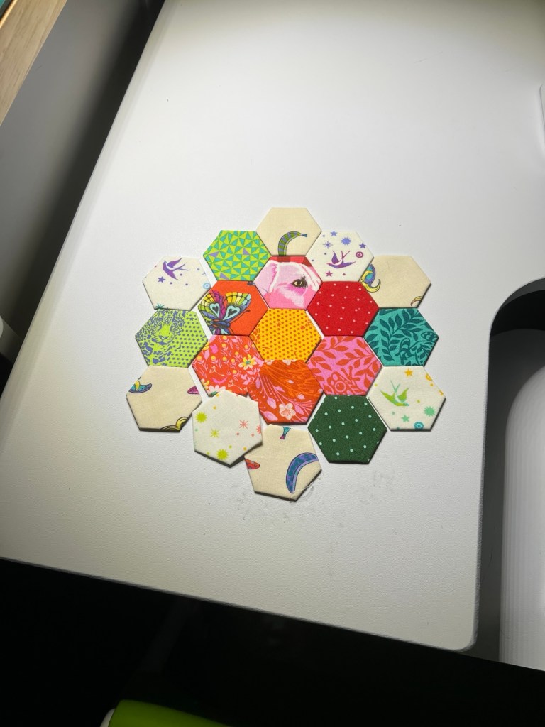 A colourful arrangement of fabric hexagons is displayed on a white table. The hexagons are placed together to form a larger hexagonal shape; a yellow reading hexagon in the centre is surrounded by 6 red reading hexagons, the surrounded by 8 white reading and 4 green reading hexagons. Each hexagon features a different fabric pattern, including florals, geometric designs, birds, moons, and dots in bright and pastel colours such as green, yellow, red, pink, and blue. Some hexagons are cream with decorative prints, while others are solid or patterned with more vibrant hues. The pieces are neatly arranged, and each fabric has been cut precisely into a hexagonal shape. The lighting highlights the textures and colours vividly.