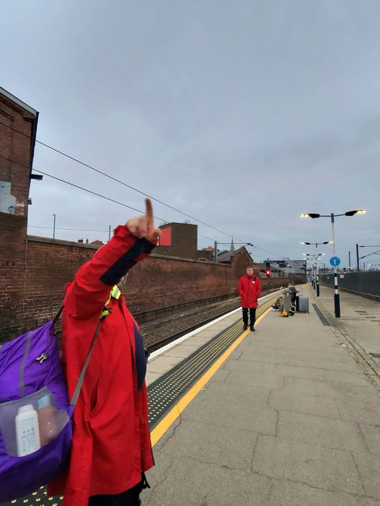 Train station platform scene. There is a person in the foreground wearing a red coat, raising their left arm, pointing with their index finger upwards. They're carrying a large purple bag with a transparent pocket that appears to have bottles inside. Further down the platform, another person, also in a red coat, is standing and looking toward the first person. The platform has a yellow tactile strip along the edge, and there are lamp posts with lights on. The sky is overcast, and there is a brick wall lining one side of the platform. Some luggage or bags are visible near the second person.